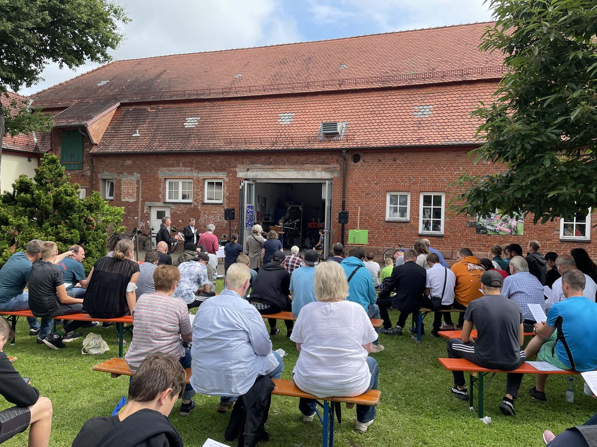 Eine Menschenmenge sitzt auf Bänken im Freien auf einem Rasen, vor einem Backsteingebäude mit Band und Rednern.