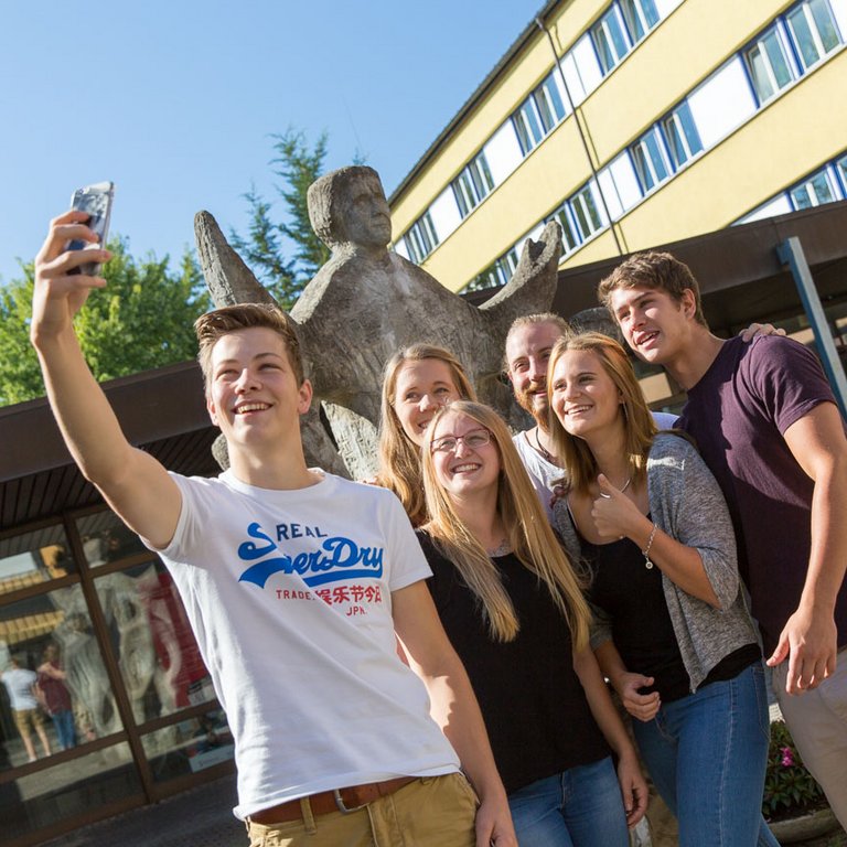 Sechs fröhliche Studierende machen ein Selfie vor einem Campusgebäude und einer Statue an einem hellen Tag.