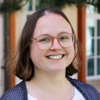 Eine lächelnde junge Frau mit braunen Haaren und Brille, die eine gemusterte Strickjacke trägt, blickt direkt in die Kamera.