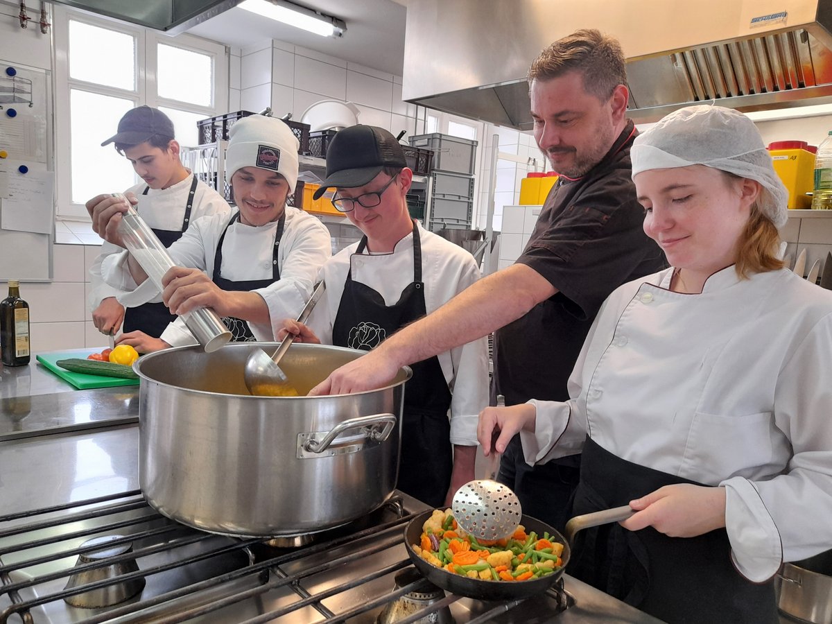 Eine Gruppe von Kochschülern und ein Lehrer kochen in einer professionellen Küche, rühren in einem Topf und braten Gemüse.