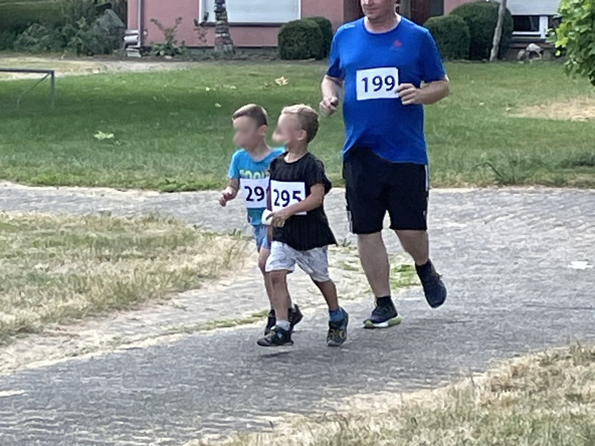 Ein Mann und zwei Jungen mit Startnummern laufen auf einem asphaltierten Weg, mit Gras, Bäumen und einem Haus im Hintergrund.