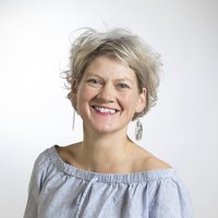 Eine lächelnde Frau mit kurzen, welligen blond-grauen Haaren und einem gestreiften schulterfreien Top blickt in die Kamera.