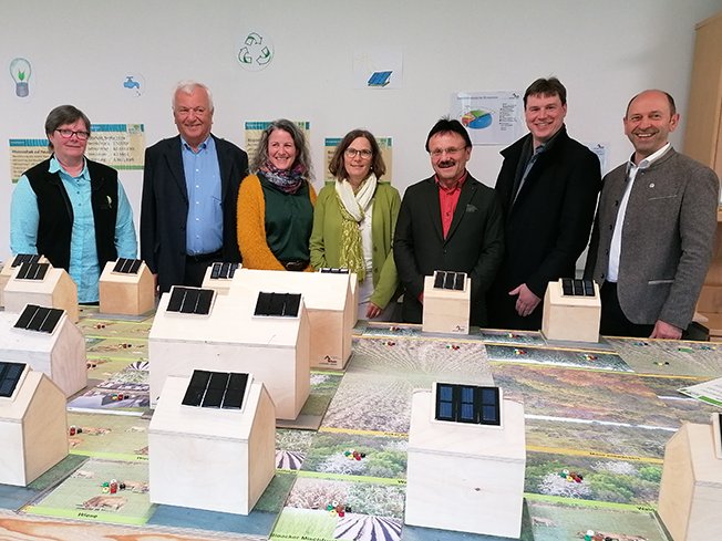 Sieben Personen hinter einem Tisch mit Modellhäusern mit Solarpaneelen, die über nachhaltige Energielösungen sprechen.