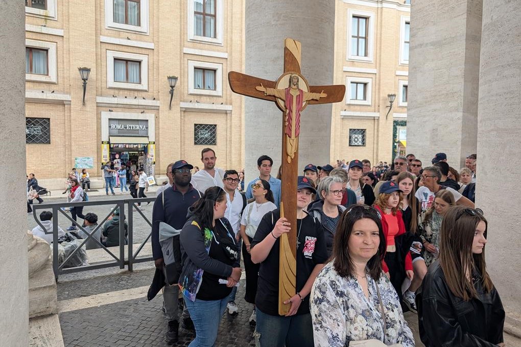 Eine Person hält ein großes Holzkruzifix in einer Menschenmenge auf einem belebten Platz mit historischen Gebäuden.