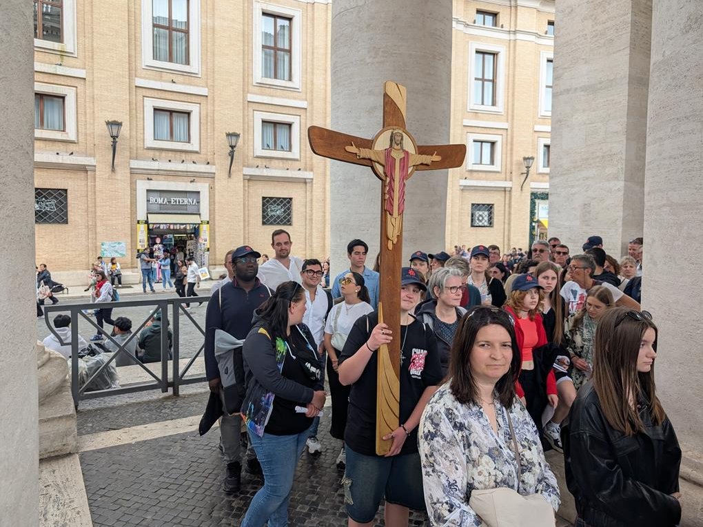 Eine Person hält ein großes Holzkruzifix in einer Menschenmenge auf einem belebten Platz mit historischen Gebäuden.