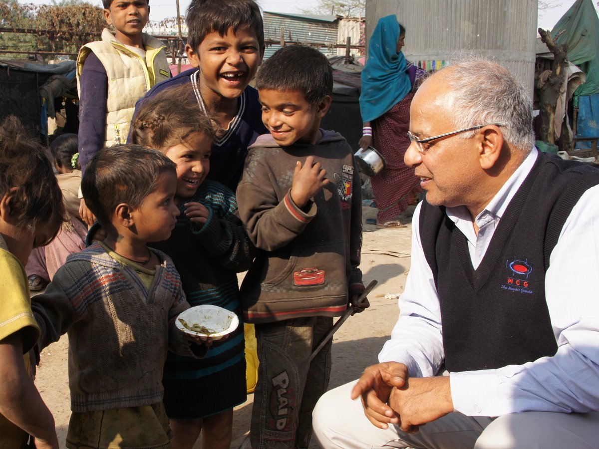 Ein älterer Mann spricht mit einer Gruppe lachender Kinder in einem informellen Siedlung, ein Kind hält eine Essensschale.