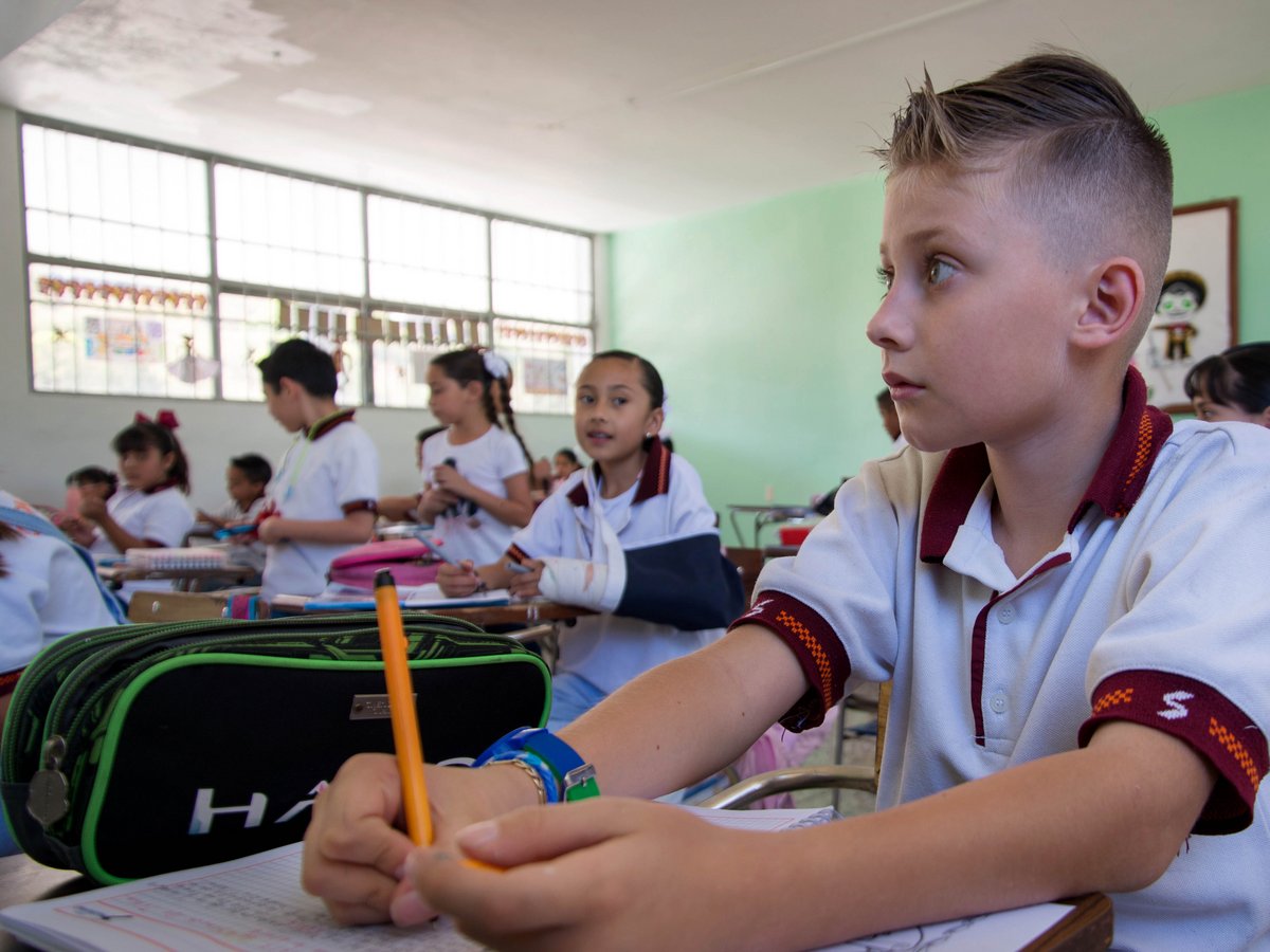 Ein Junge mit gestylten Haaren sitzt an einem Schreibtisch in einem belebten Klassenzimmer, einen Stift haltend. Andere Schüler sind sichtbar.