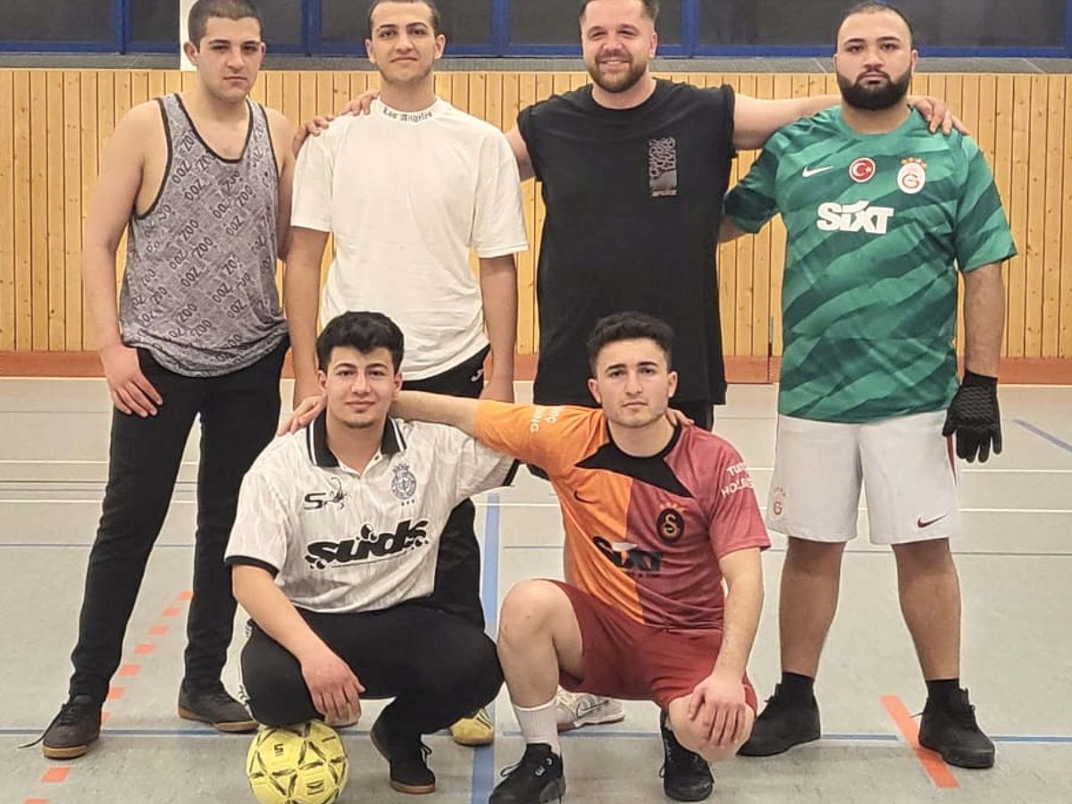 Sechs junge Männer, einige in Fußballtrikots, posieren mit einem Ball in einer Sporthalle. Drei stehen, drei hocken.