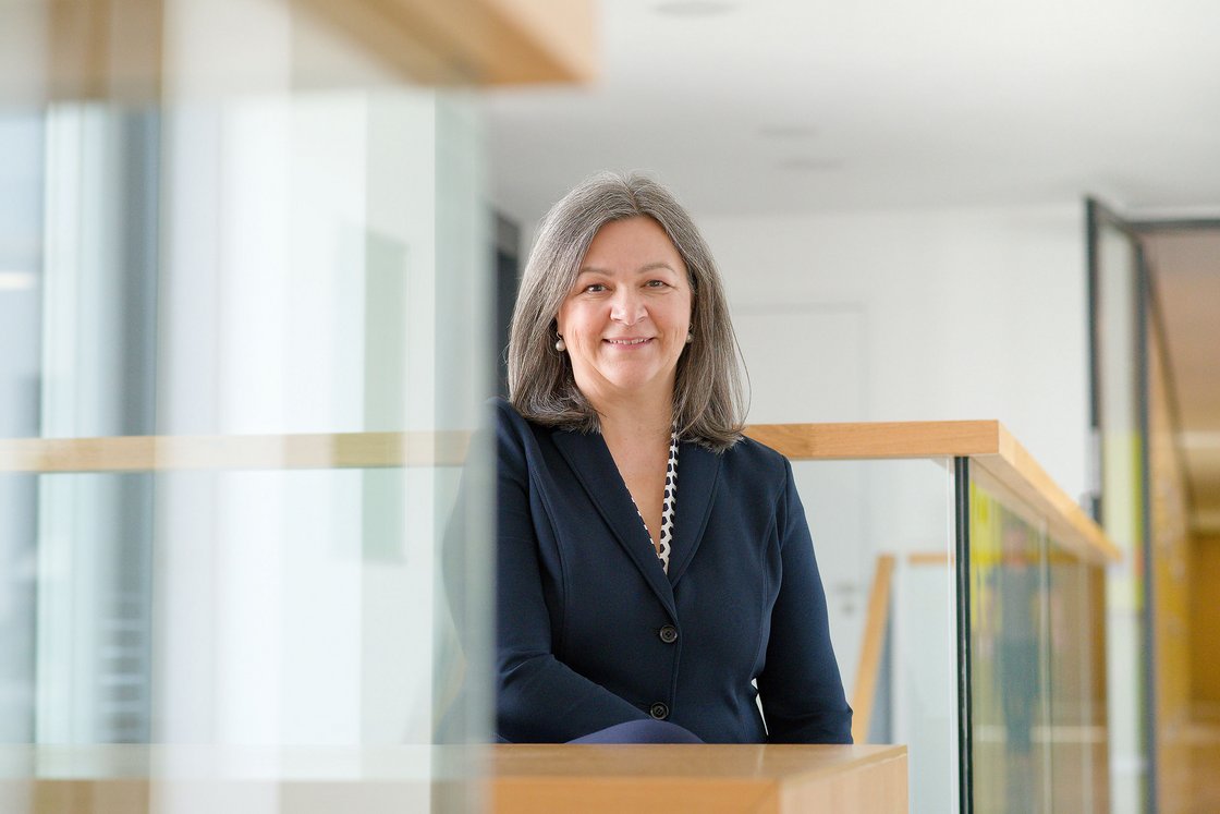 Lächelnde Frau mit grauen Haaren und dunklem Blazer sitzt hinter einer Glas- und Holzbrüstung in einem modernen Interieur.