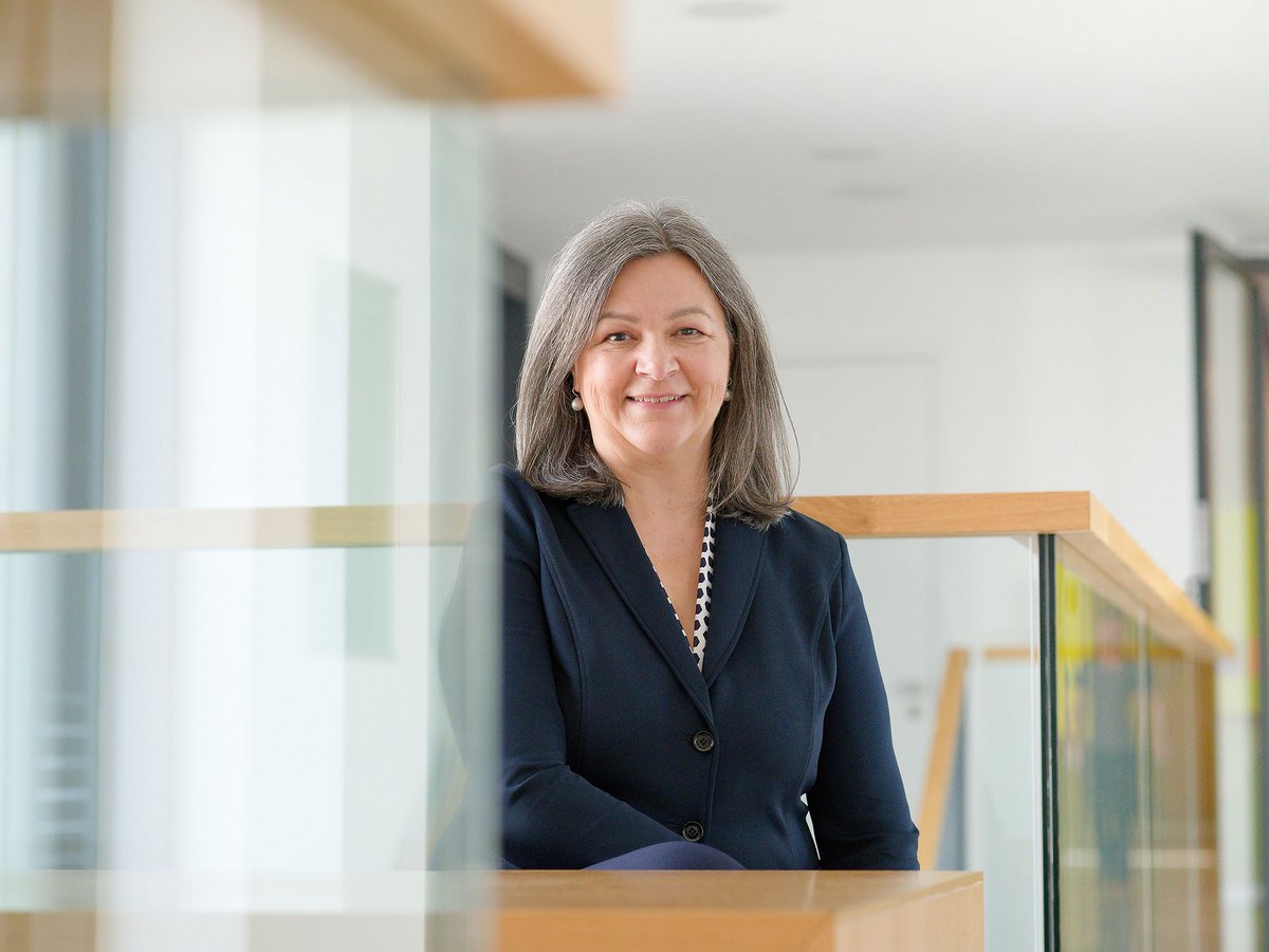 Lächelnde Frau mit grauen Haaren und dunklem Blazer sitzt hinter einer Glas- und Holzbrüstung in einem modernen Interieur.