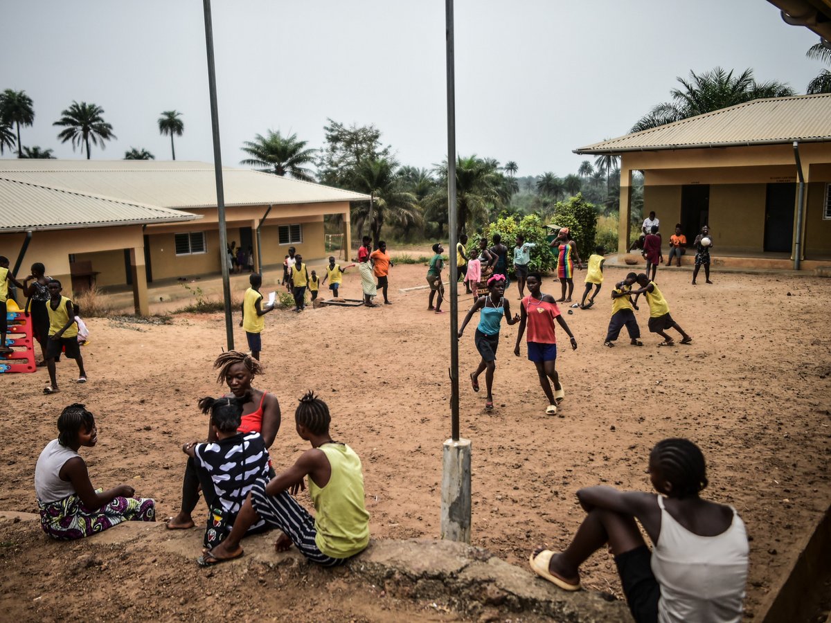 Kinder spielen und unterhalten sich auf einem staubigen Schulhof mit Gebäuden und Palmen im Hintergrund.