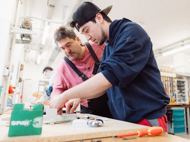 Zwei Männer, ein Jüngerer mit Kappe und ein Älterer, verdrahten Elektrik an einer Werkbank in heller Werkstatt.