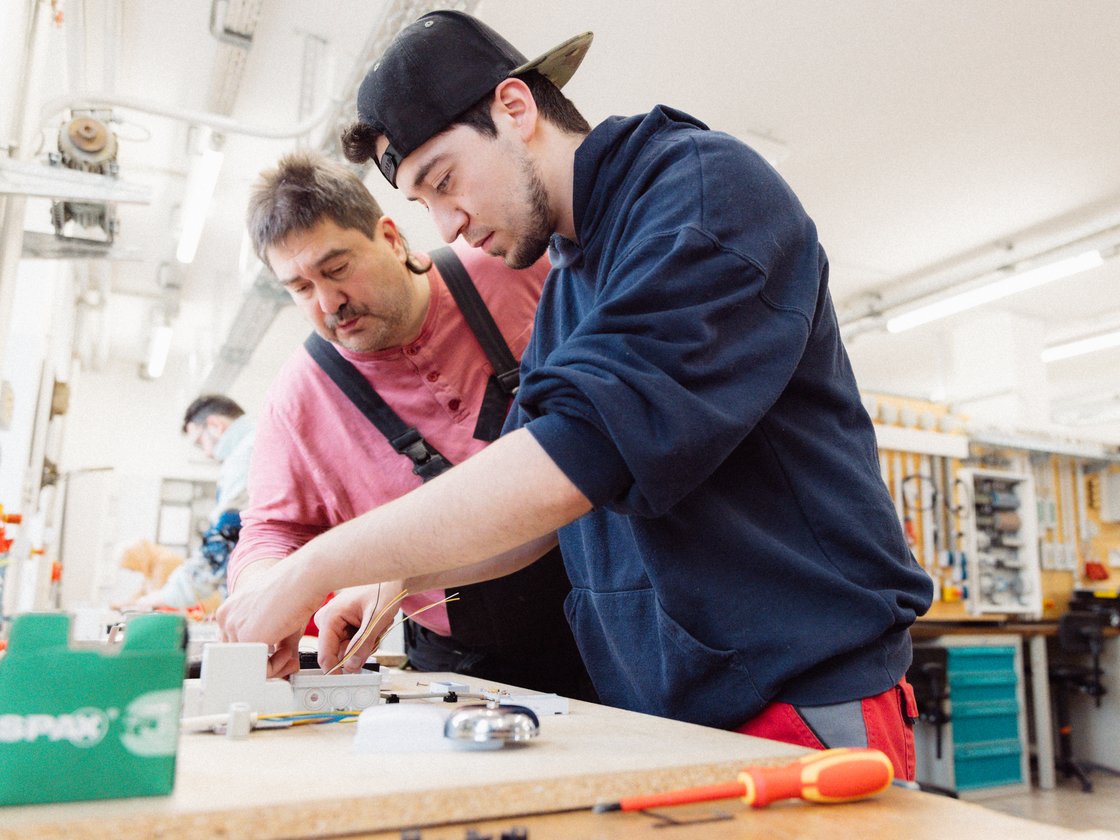 Zwei Männer, ein Jüngerer mit Kappe und ein Älterer, verdrahten Elektrik an einer Werkbank in heller Werkstatt.