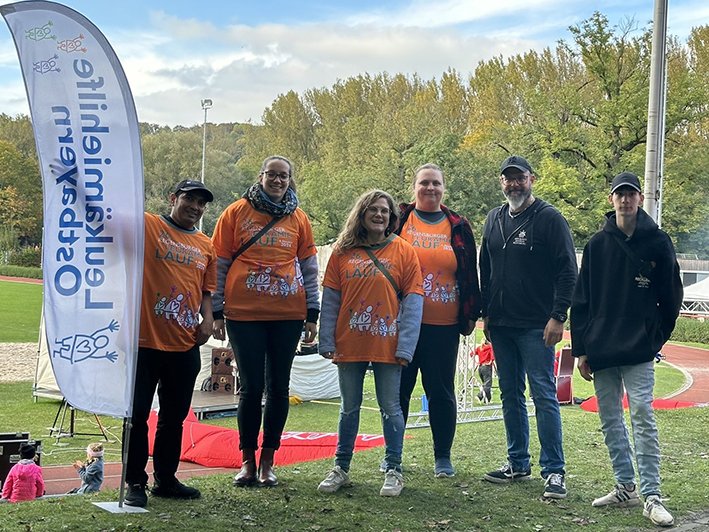 Sechs Personen in orangefarbenen T-Shirts des Hildburghäuser Grenzlandlaufs, posieren auf einer Sportanlage neben einem Banner.