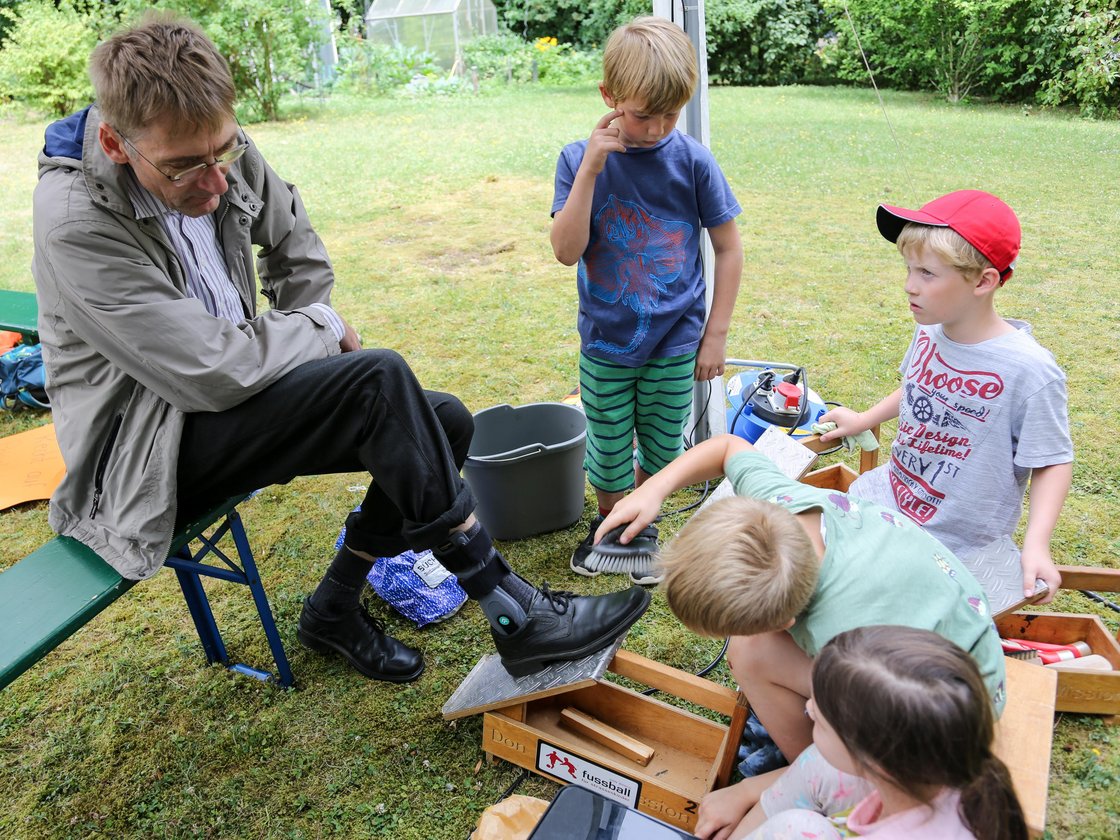 Ein Mann sitzt auf einer Bank, während mehrere Kinder ihm draußen die Schuhe putzen, mit einem Gewächshaus im Hintergrund.