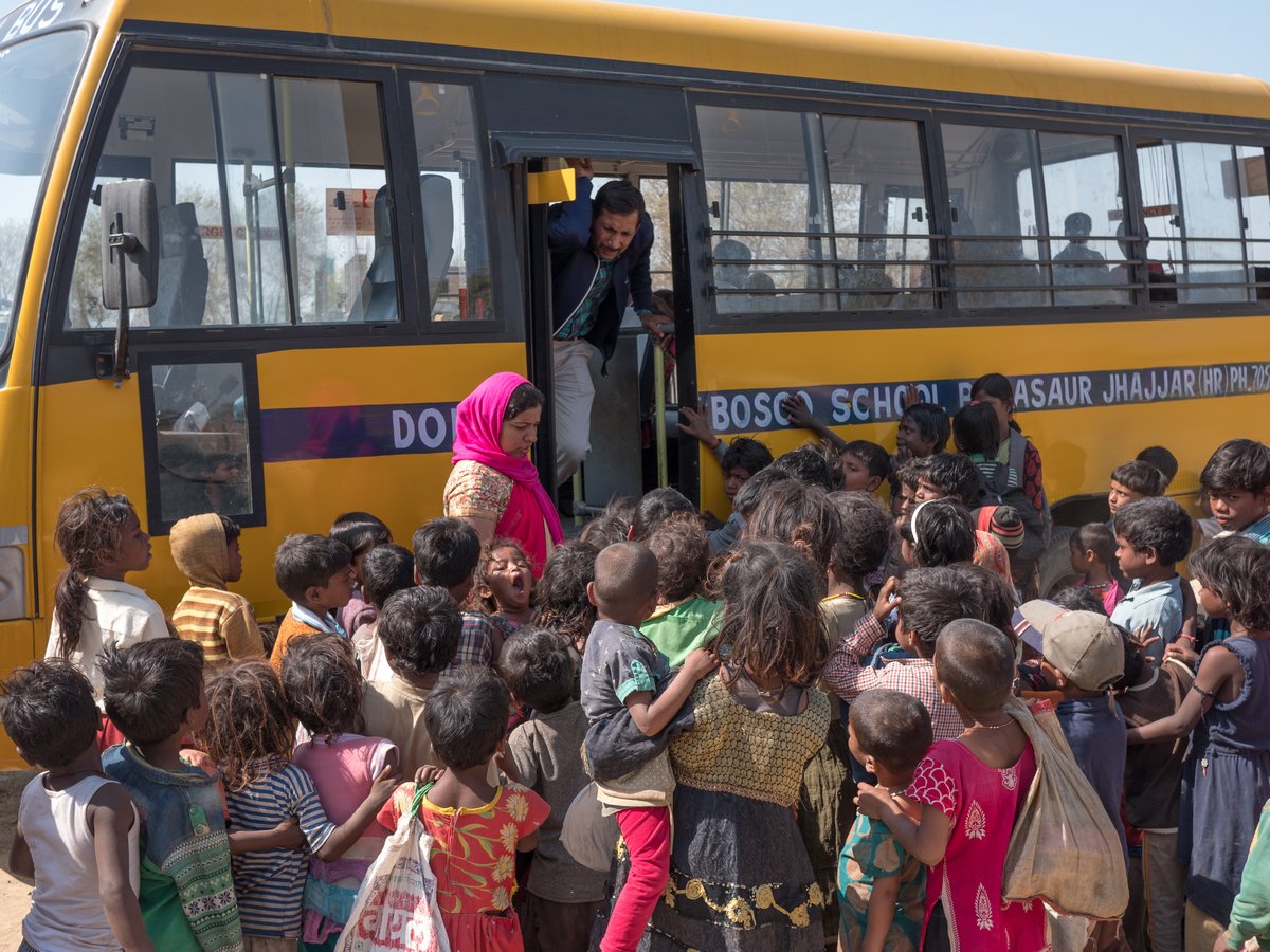 Ein Mann und eine Frau interagieren mit einer großen Gruppe von Kindern, die sich um einen gelben "Don Bosco School" Bus versammelt hat.