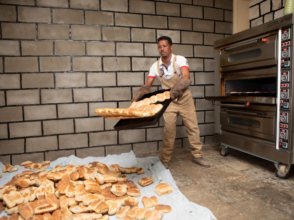 Mann in Schürze und Ofenhandschuhen nimmt Blech mit goldbraunen Broten aus Backofen, neben großem Brothaufen.