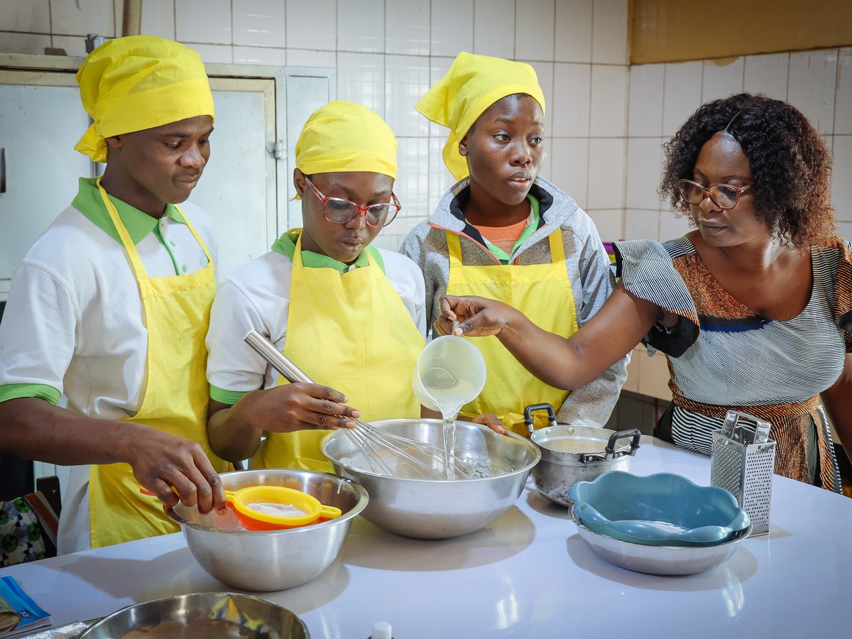 Vier Personen, darunter eine Ausbilderin und drei Schüler in gelben Schürzen, bereiten Speisen in einer Küche zu.