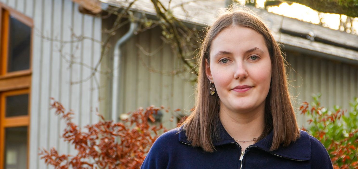 Eine junge Frau im blauen Pullover lächelt leicht, steht draußen vor einem Gebäude mit roten Büschen und einem moosigen Ast.