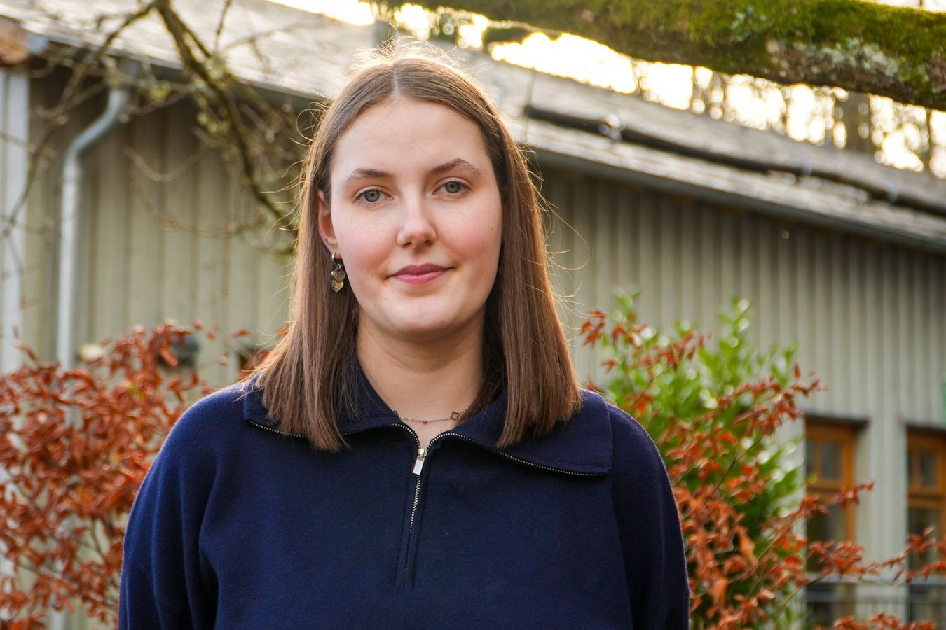 Eine junge Frau im blauen Pullover lächelt leicht, steht draußen vor einem Gebäude mit roten Büschen und einem moosigen Ast.