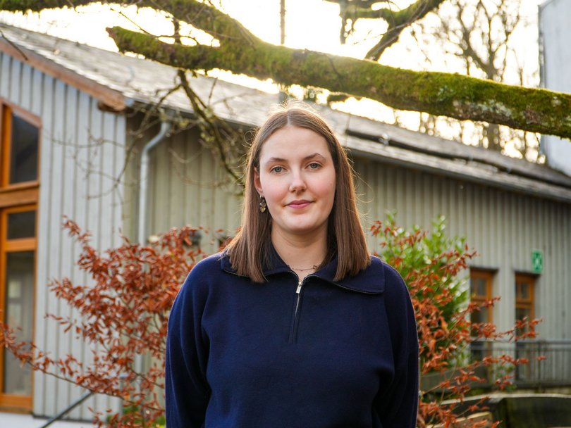 Eine junge Frau im blauen Pullover lächelt leicht, steht draußen vor einem Gebäude mit roten Büschen und einem moosigen Ast.