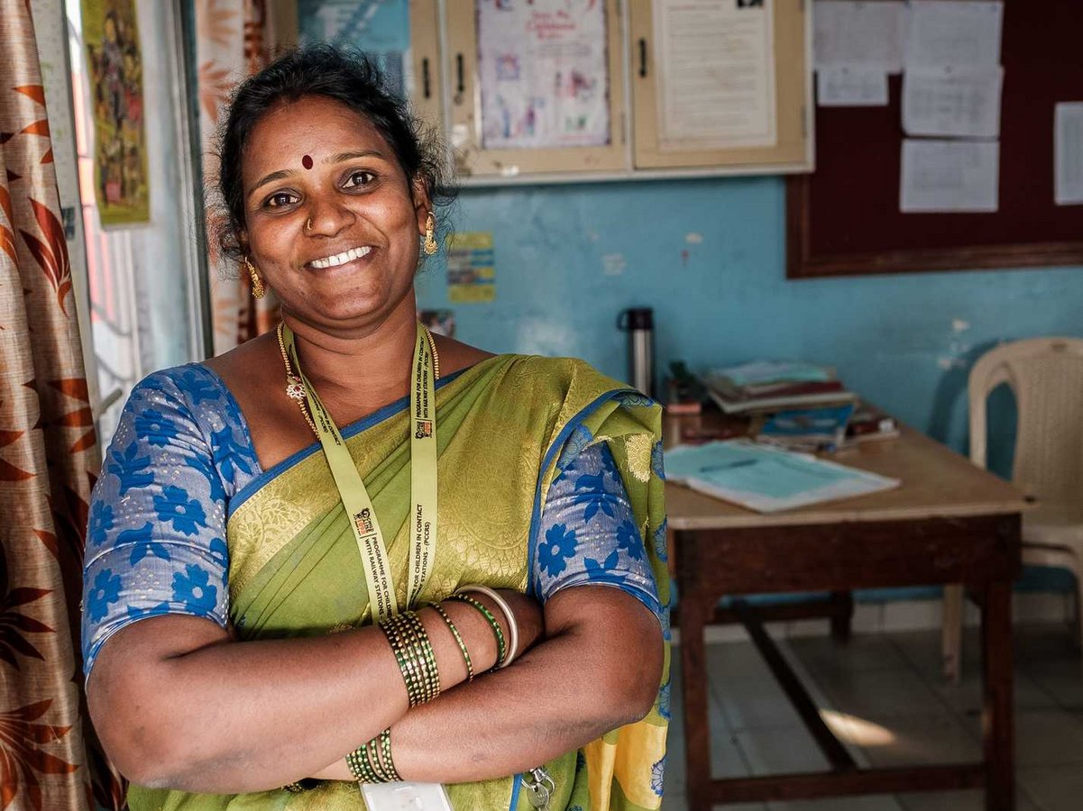 Eine lächelnde indische Frau in grünem Sari und blauer Bluse steht mit verschränkten Armen in einem Büro mit 'Child Help Desk' Schild.