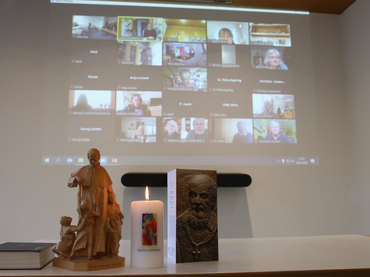 Ein Beamer zeigt eine Videokonferenz, davor auf einem Tisch eine Holzstatue, eine brennende Kerze und ein Buch.