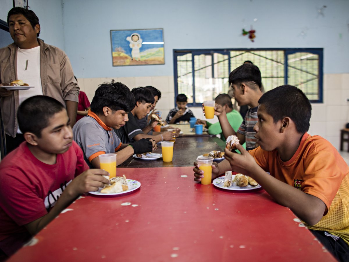 Eine Gruppe junger Männer und Jungen sitzt an einem roten Tisch, isst Gebäck und trinkt Orangensaft, während ein erwachsener Mann beaufsichtigt.