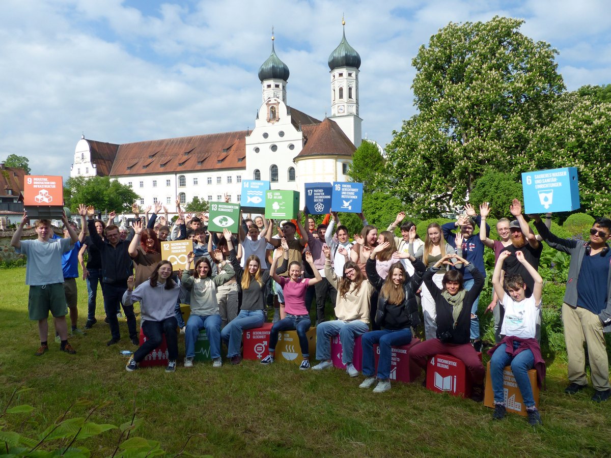 Eine Gruppe junger Leute posiert lachend mit SDGs-Schildern und -Würfeln vor einem historischen Klostergebäude mit Zwiebeltürmen.
