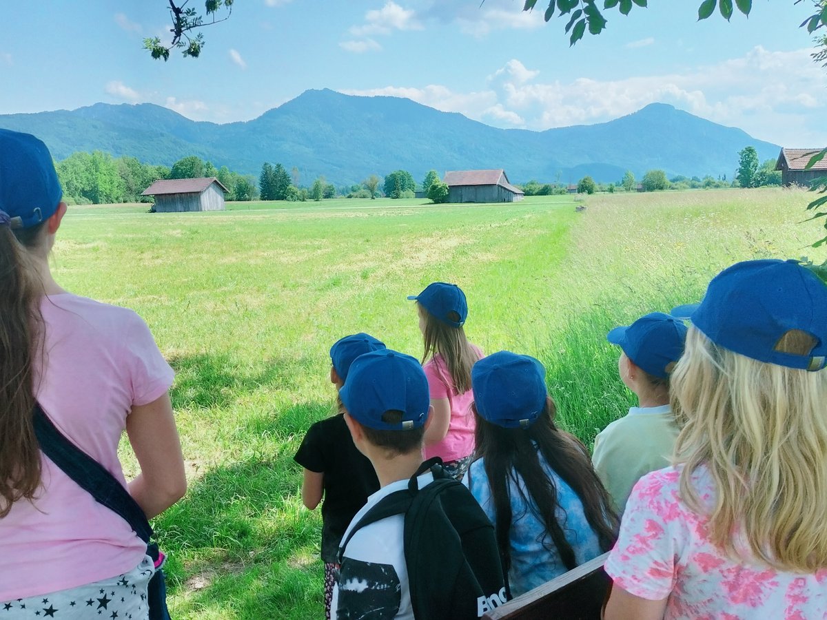 Eine Gruppe Kinder mit blauen Kappen blickt auf eine weite Wiese mit Holzhütten und Bergen unter blauem Himmel.