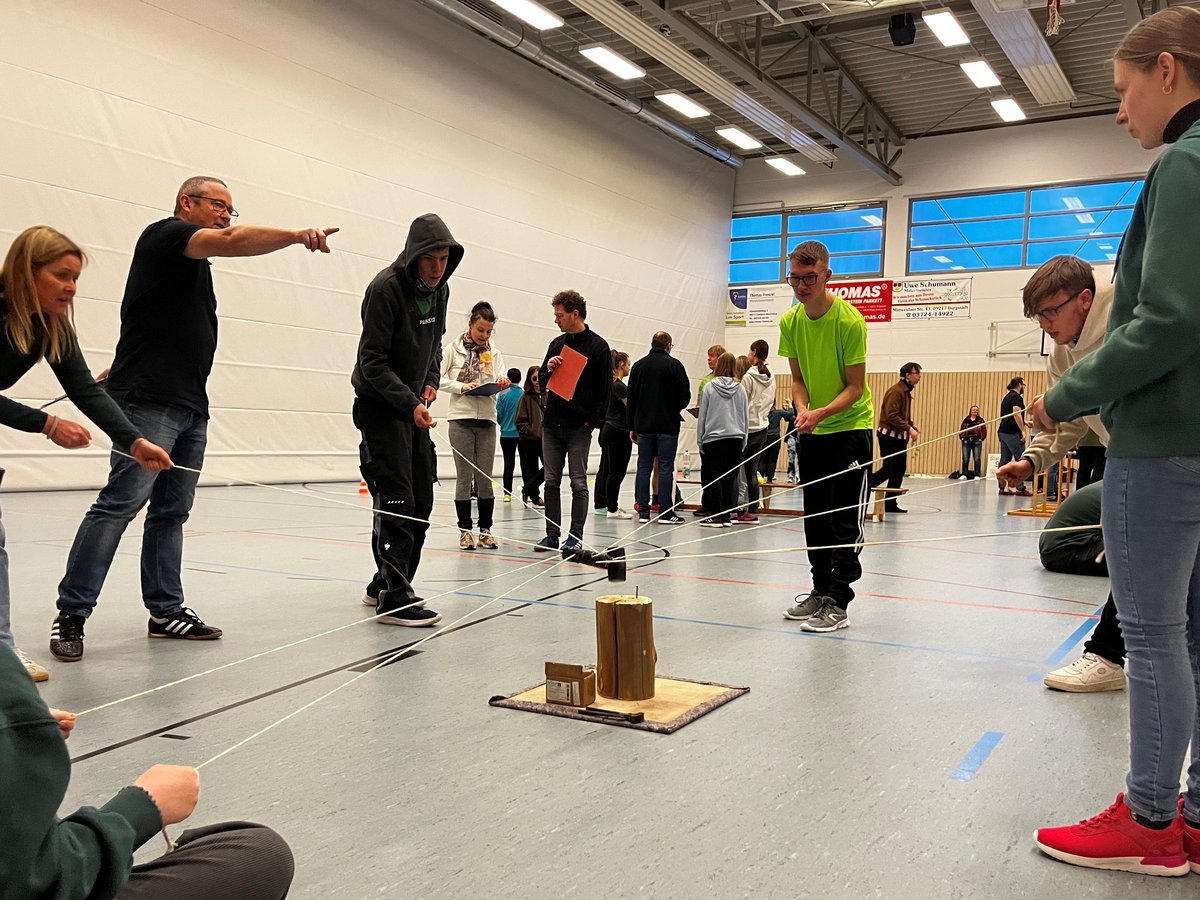 Jugendliche und Erwachsene in einer Turnhalle spielen ein Kooperationsspiel mit Seilen und einem Holzobjekt.