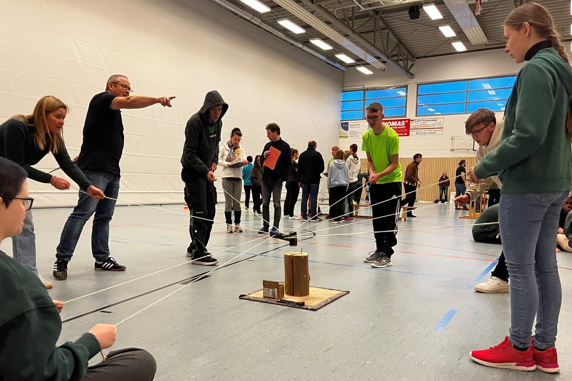Jugendliche und Erwachsene in einer Turnhalle spielen ein Kooperationsspiel mit Seilen und einem Holzobjekt.