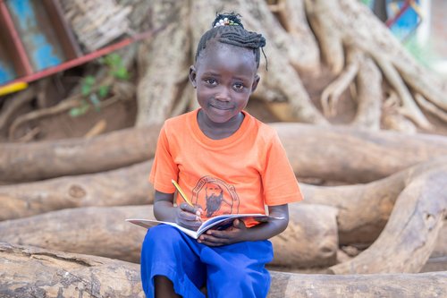 Lächelndes junges afrikanisches Mädchen sitzt draußen, schreibt in Notizbuch mit Bleistift. Trägt orangefarbenes Hemd und blaue Hose.