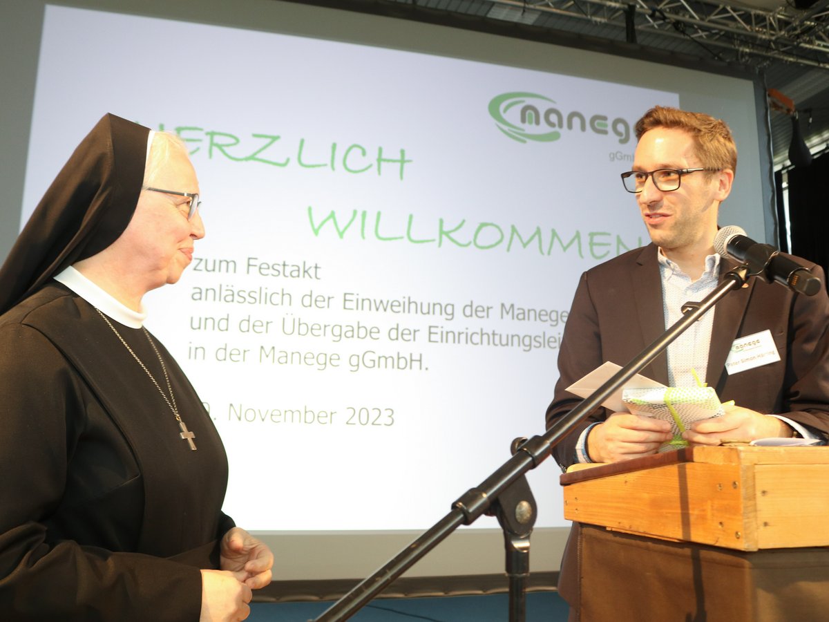 Eine Nonne und ein Mann im Anzug begrüßen sich an einem Pult. Er hält ein Geschenk. Hintergrund: 'Willkommen' und 'Manege gGmbH'.