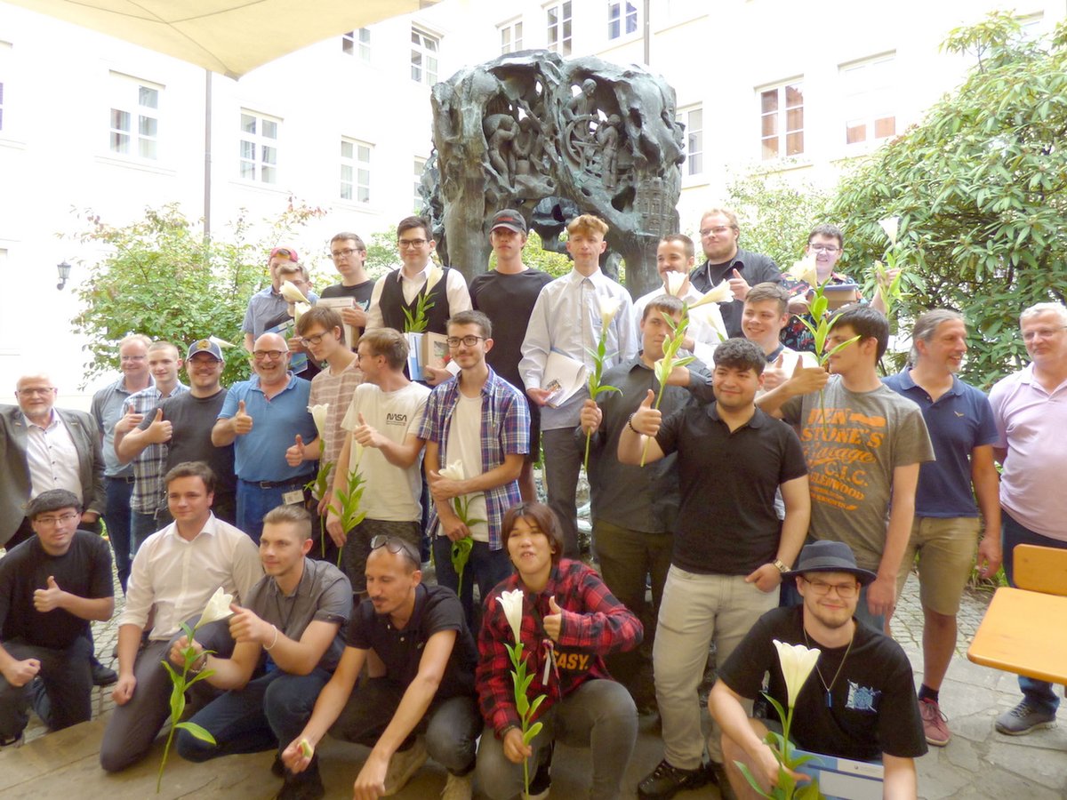 Eine große Gruppe Männer, einige mit weißen Lilien, geben Daumen hoch im Freien vor einer dunklen Skulptur.
