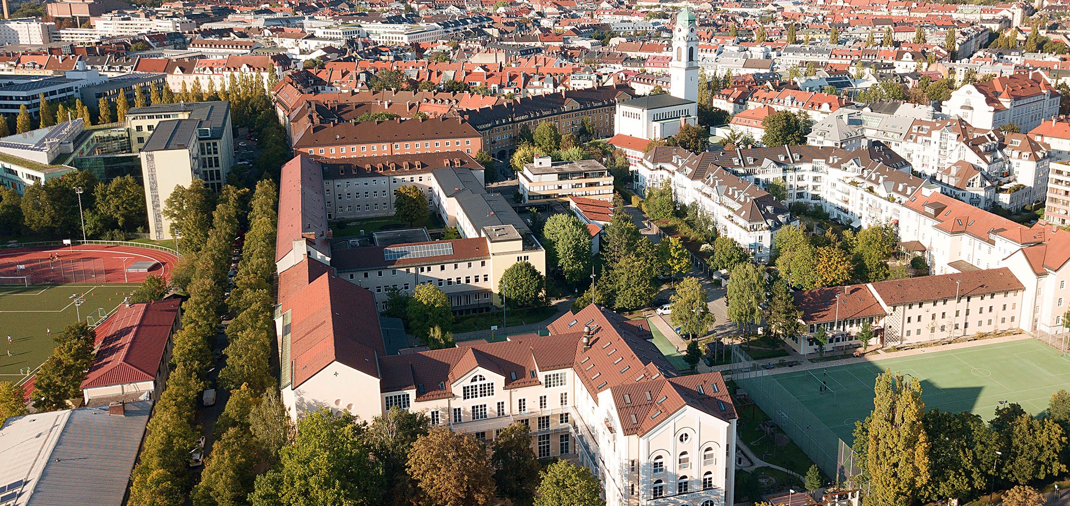 Vogelperspektive auf ein Ensemble von Gebäuden mit roten Dächer, Sportplatz und Begrünung.