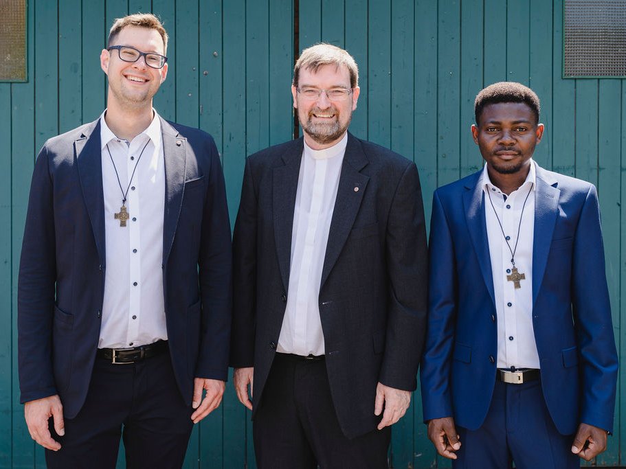 Drei Geistliche, zwei mit Kruzifixen, einer mit Priesterkragen, lächeln vor einer türkisfarbenen Holztür.