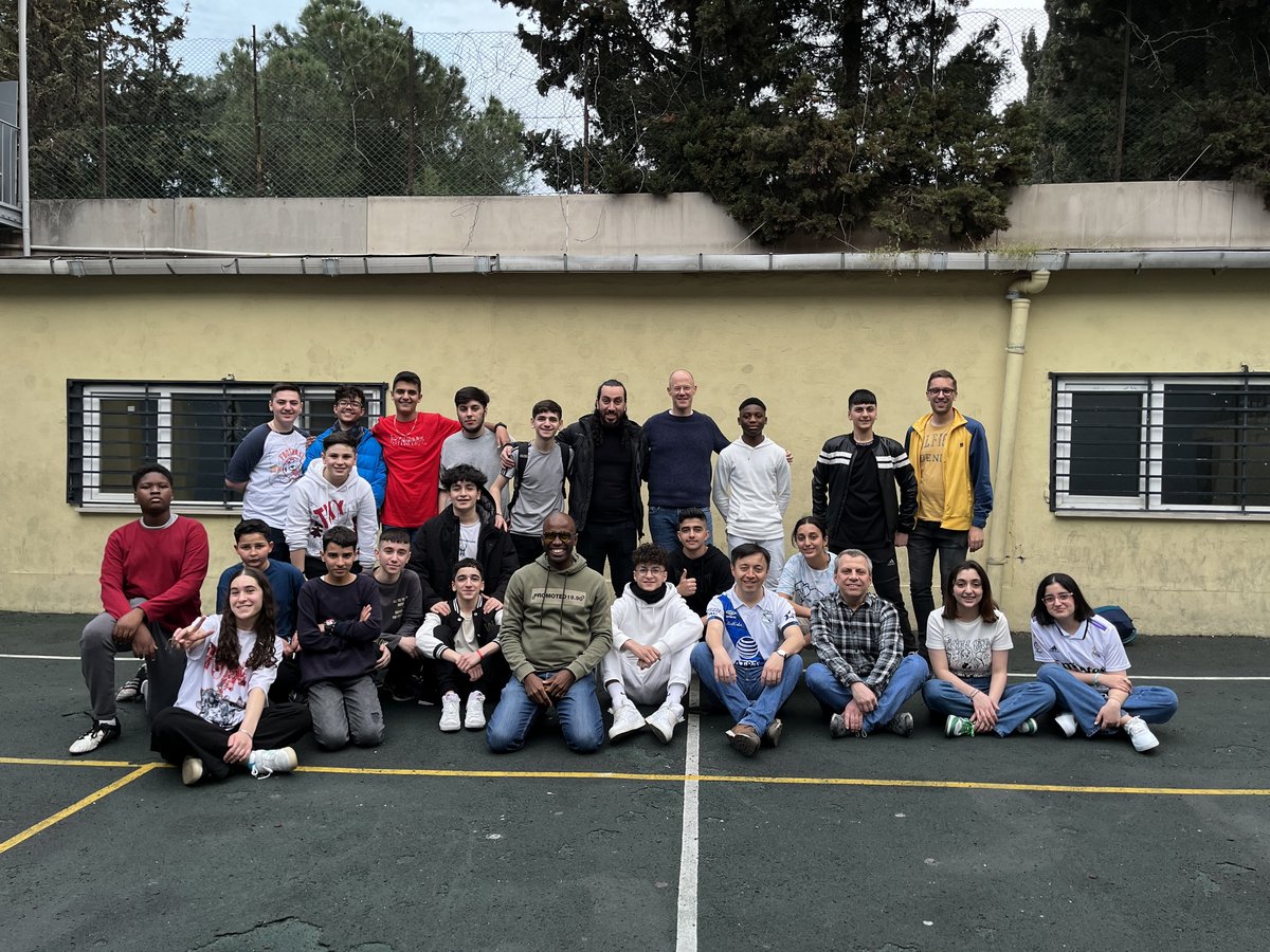 Eine vielfältige Gruppe von Schülern und Erwachsenen, hauptsächlich Männer, posiert draußen auf einem Schulhof mit Gebäude und Bäumen.
