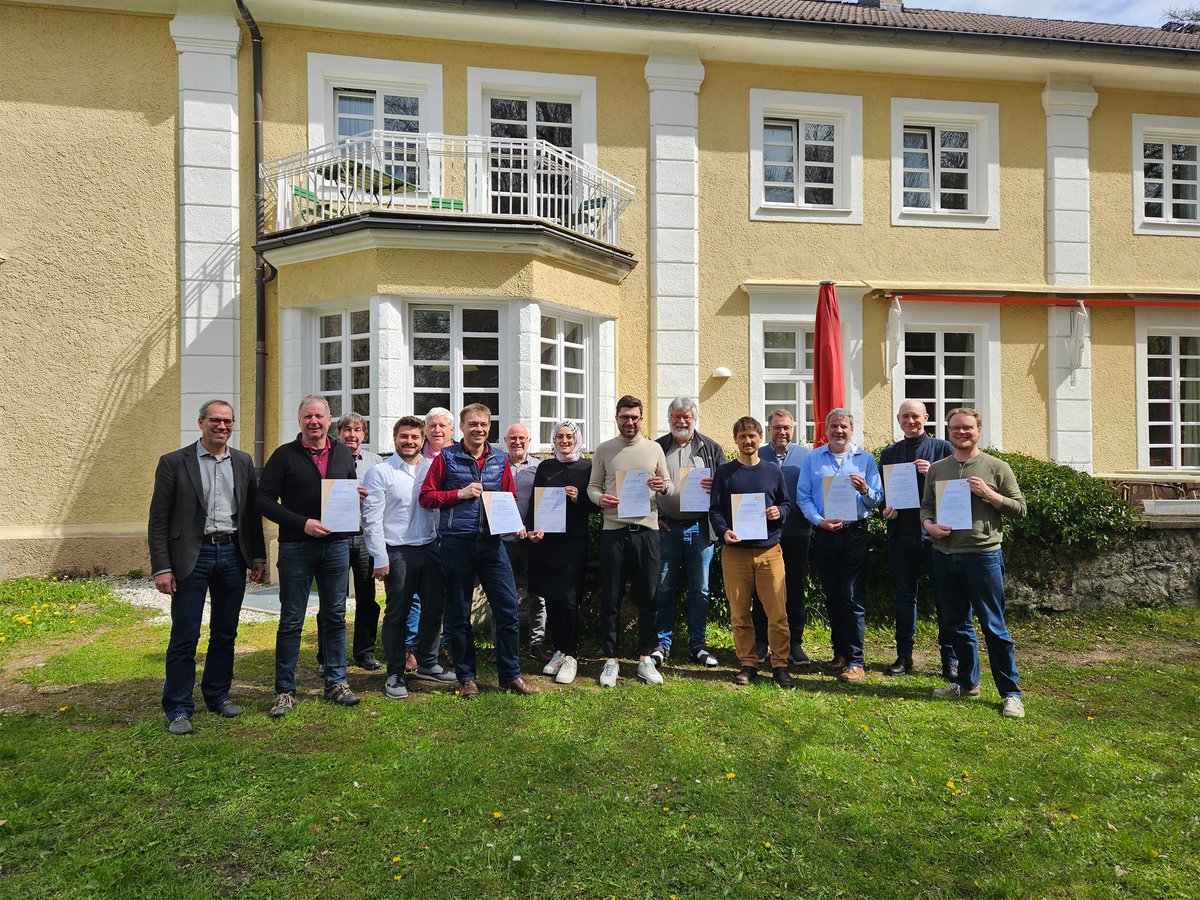 Eine Gruppe von Personen hält Zertifikate auf einem Rasen vor einem großen gelben Gebäude mit weißen Verzierungen.