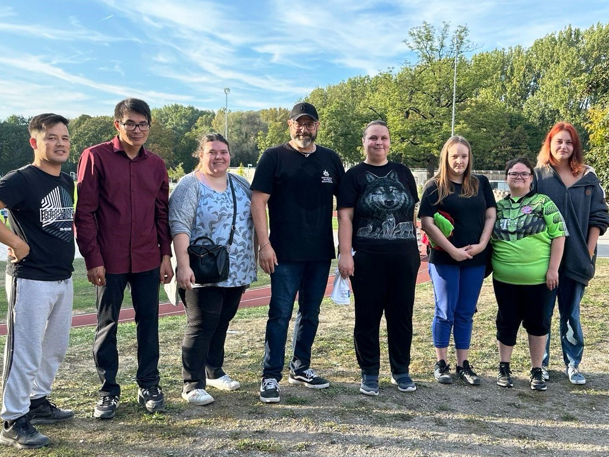 Gruppenfoto von acht Erwachsenen, fünf Frauen und drei Männern, die draußen auf einem Grasfeld mit Bäumen im Hintergrund stehen.