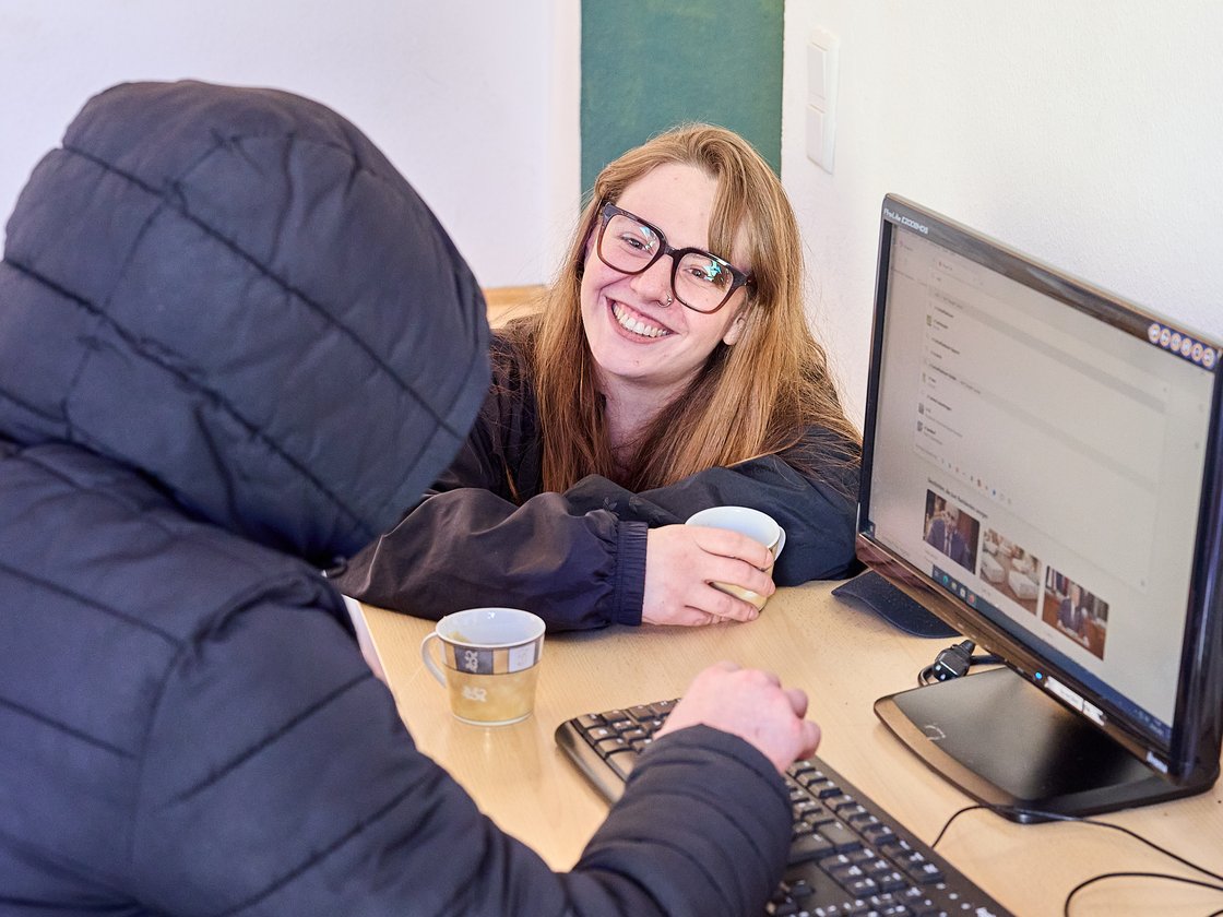 Zwei Personen an einem Schreibtisch, eine von hinten tippt auf einer Tastatur, eine lächelnde Frau mit Brille und Tasse blickt zu ihnen.