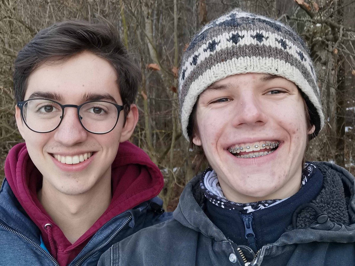 Zwei lächelnde junge Männer, einer mit Brille, der andere mit Zahnspange und Mütze, in einem Winterwald mit Schnee.