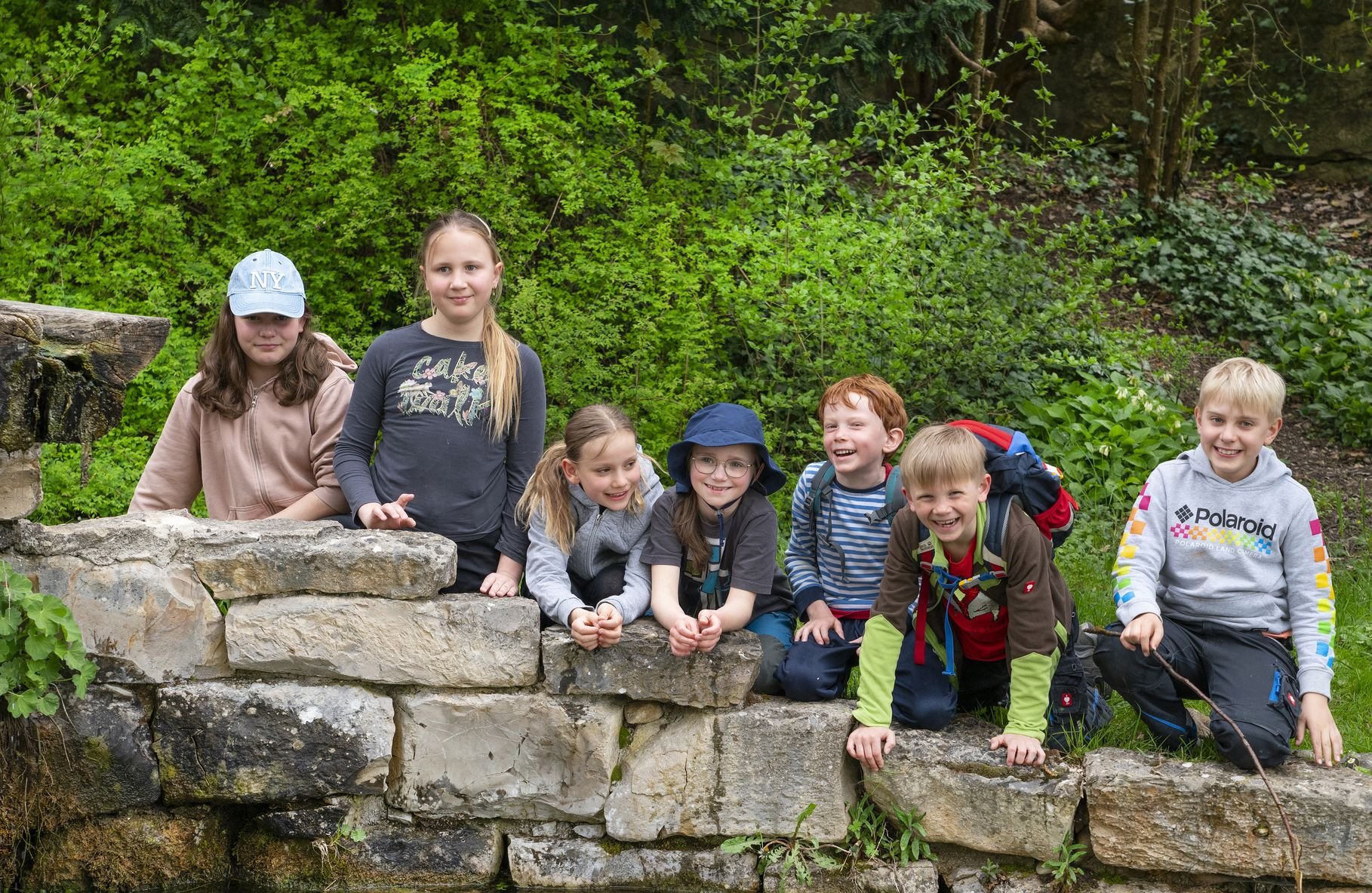 Sieben fröhliche Kinder, vier Mädchen und drei Jungen, lächeln beim Posieren an einer Steinmauer neben einem Teich in der Natur.