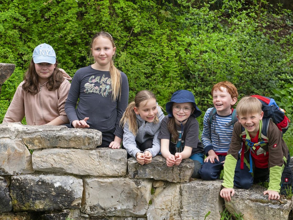 Sieben fröhliche Kinder, vier Mädchen und drei Jungen, lächeln beim Posieren an einer Steinmauer neben einem Teich in der Natur.
