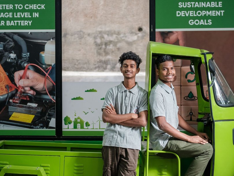 Zwei lächelnde junge Männer posieren vor einem hellgrünen Dreirad und Postern über Technik und Nachhaltigkeit.