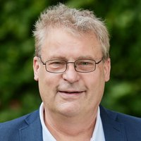 Porträt eines lächelnden Mannes mit Brille, hellem Haar, weißem Poloshirt und blauem Sakko vor grünem Hintergrund.