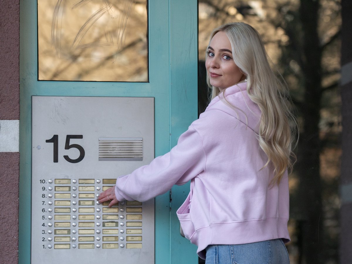 Blonde Frau in pinkfarbenem Hoodie drückt Klingelknopf an Gegensprechanlage mit Nr. 15, blickt zurück.