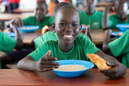 Ein lächelnder schwarzer Junge in grünem T-Shirt hält ein frittiertes Gebäck und eine Schale Essen an einem Gemeinschaftstisch mit anderen Kindern.