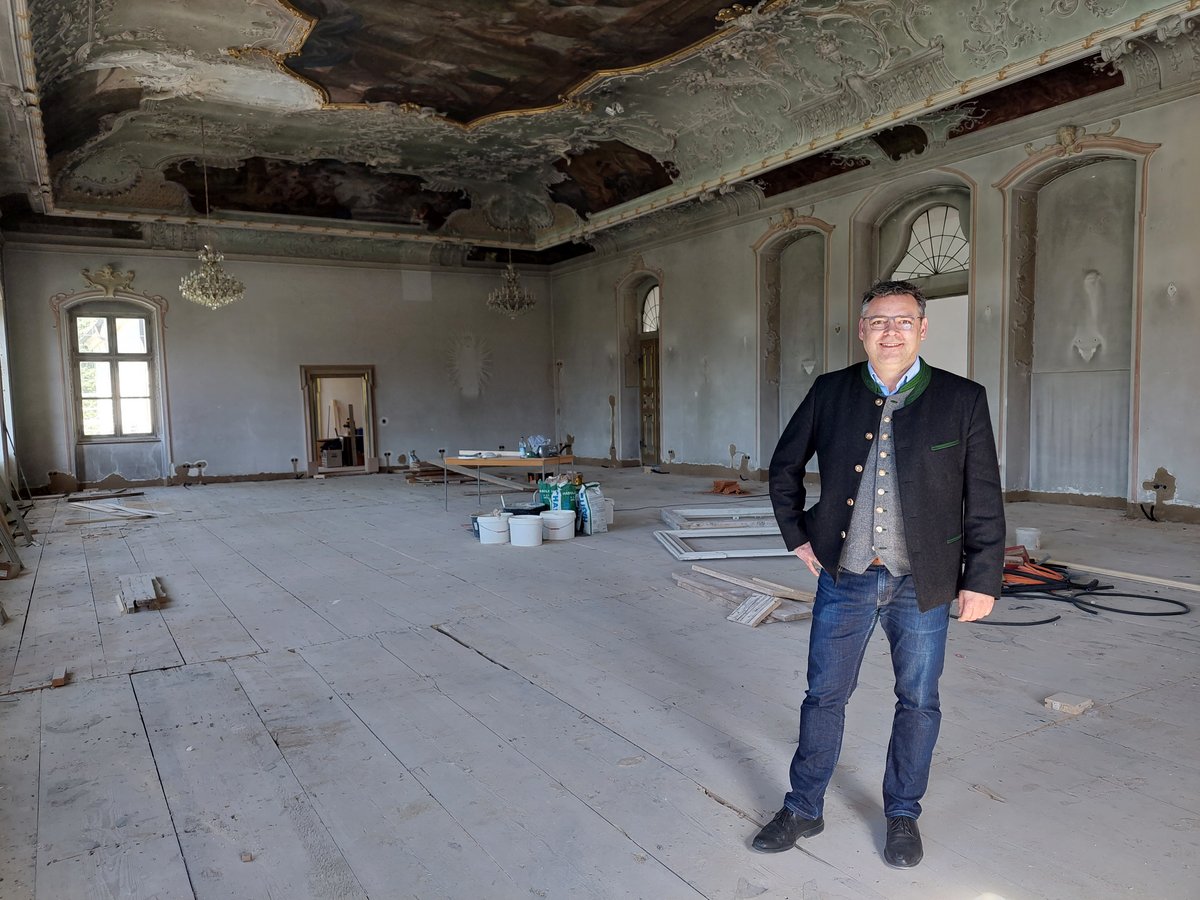 Lächelnder Mann in Trachtenjacke in großem, altem Raum mit verzierter, beschädigter Decke und Holzfußboden, wird renoviert.