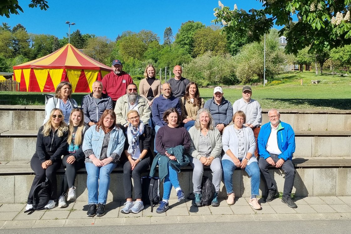 Eine Gruppe Erwachsener sitzt auf Stufen im Freien vor einem rot-gelben Zirkuszelt und Bäumen.